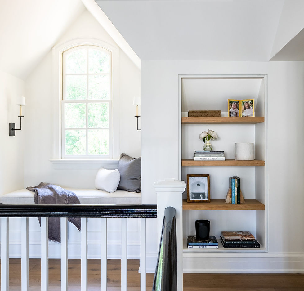 Cozy nook with bench, pillows, and a throw by a window. Nearby shelves hold books, decor, and framed photos. Bright, serene atmosphere.