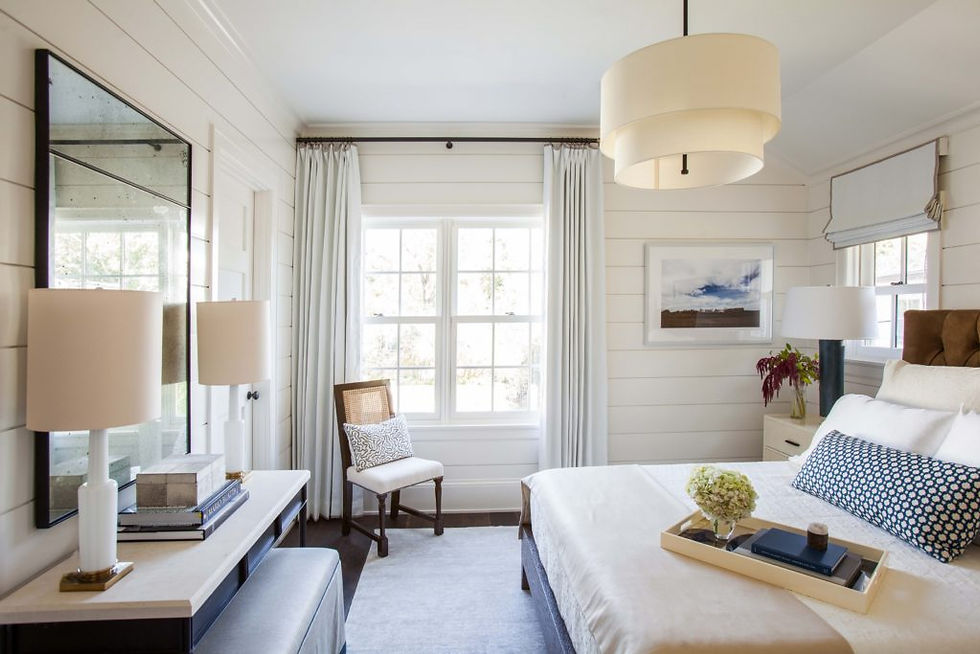 Bright bedroom with white shiplap walls, a large window, and a beige bed. Lamp-lit nightstands and a chair add cozy touches.