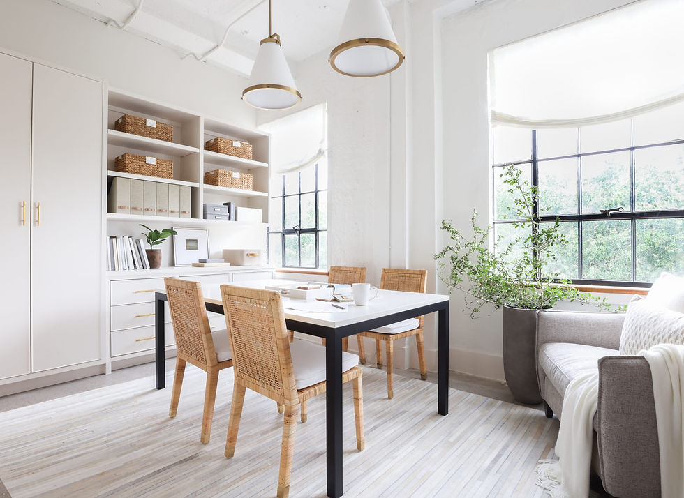 Bright dining room with a white table, wicker chairs, and a large window. Shelves with baskets and books, plants, and a cozy gray sofa.