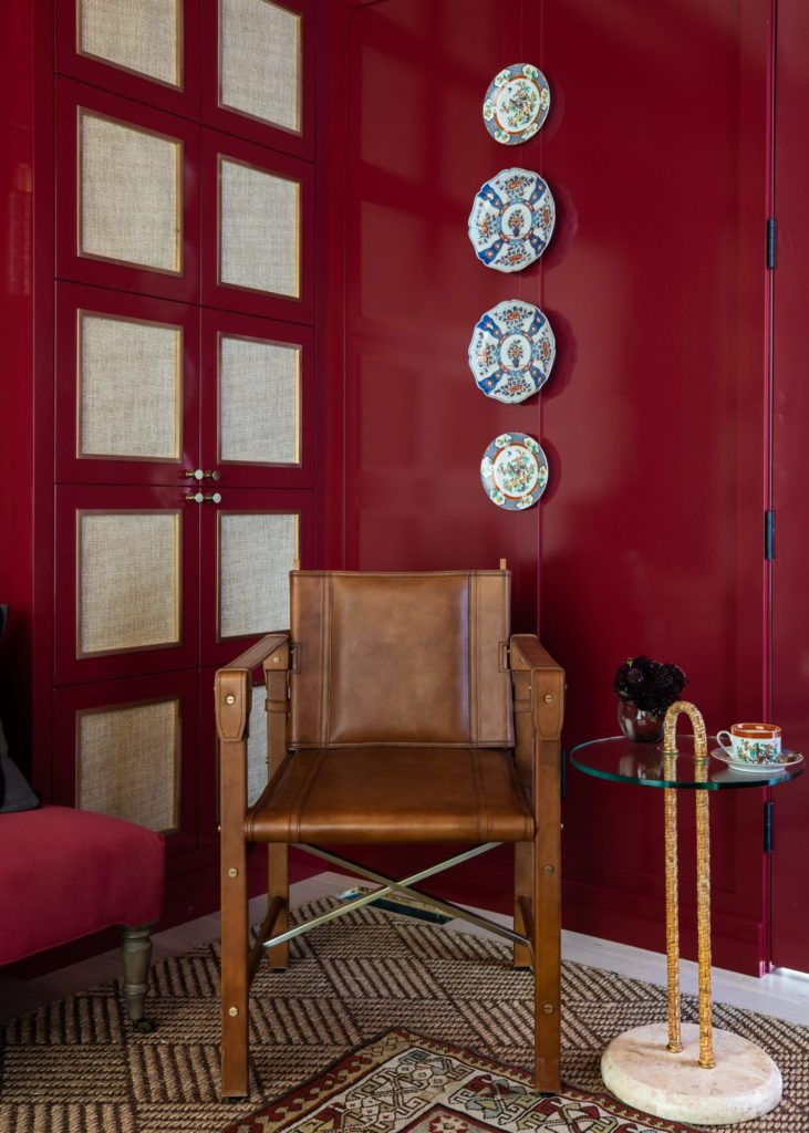 Brown leather chair in a red room with patterned plates on the wall. A glass table with flowers and a cup adds elegance. Cozy atmosphere.