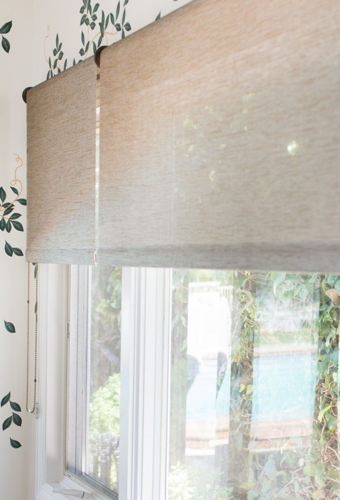 Beige window shades partially drawn, with leafy wallpaper on walls. Outside, lush greenery surrounds a visible pool area, creating a serene mood.