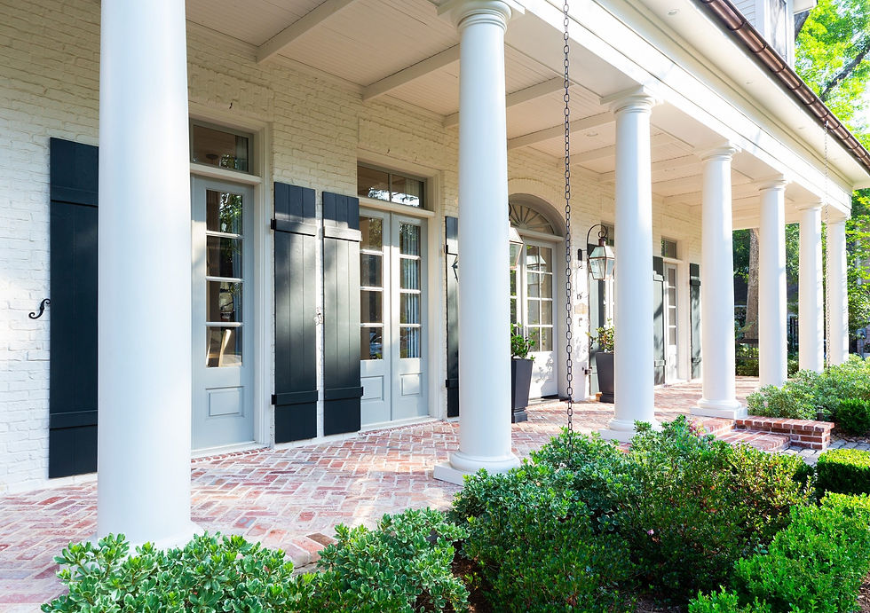 White colonial porch with tall columns, dark shutters, lanterns, and brick flooring. Green shrubs line the entrance. Bright, sunny mood.