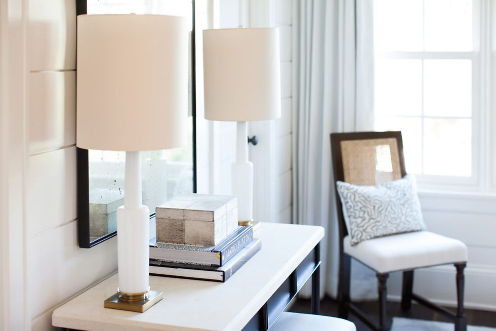 A cozy room with a desk, a white lamp, books, and a decorative box. A chair with a patterned pillow is by a window with light curtains.