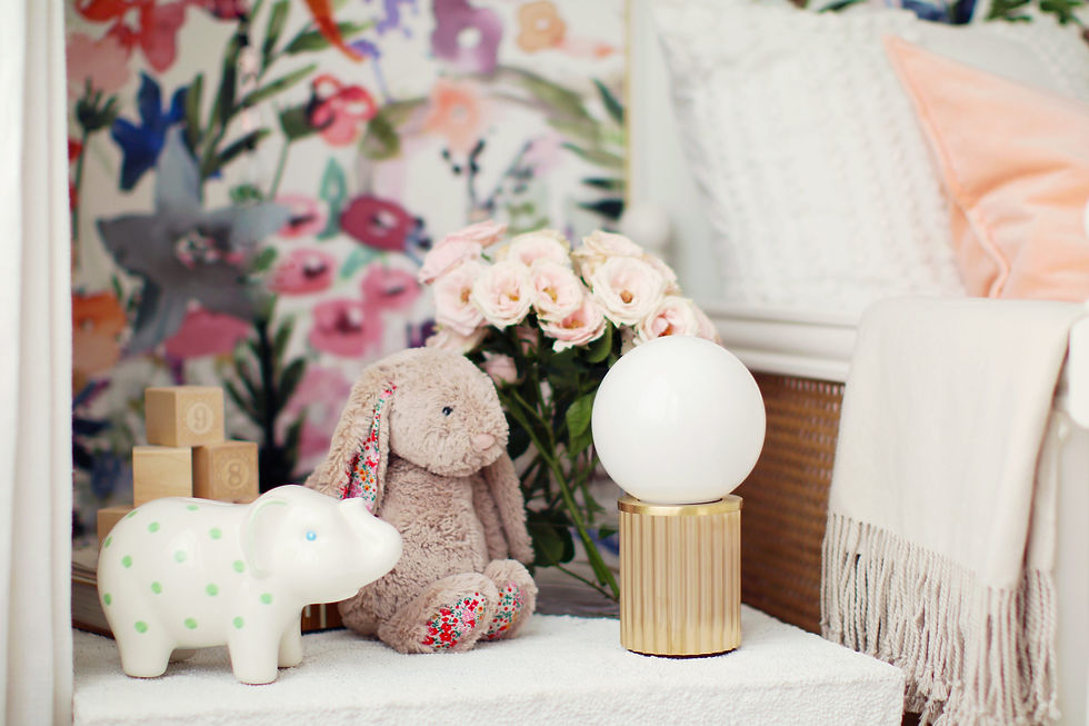 Stuffed bunny with floral ears, white ceramic hippo with green dots, and a round lamp on a table. Floral wallpaper, pink roses. Cozy vibe.