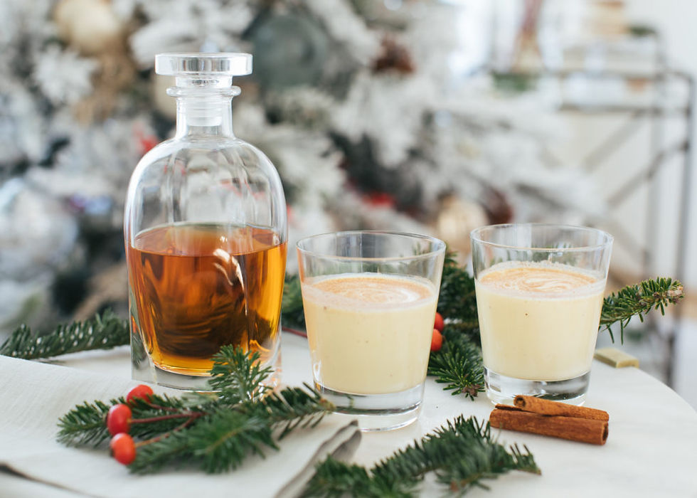 Decanter of amber liquid and two glasses of eggnog on a table with pine branches and cinnamon sticks. Festive, cozy background.