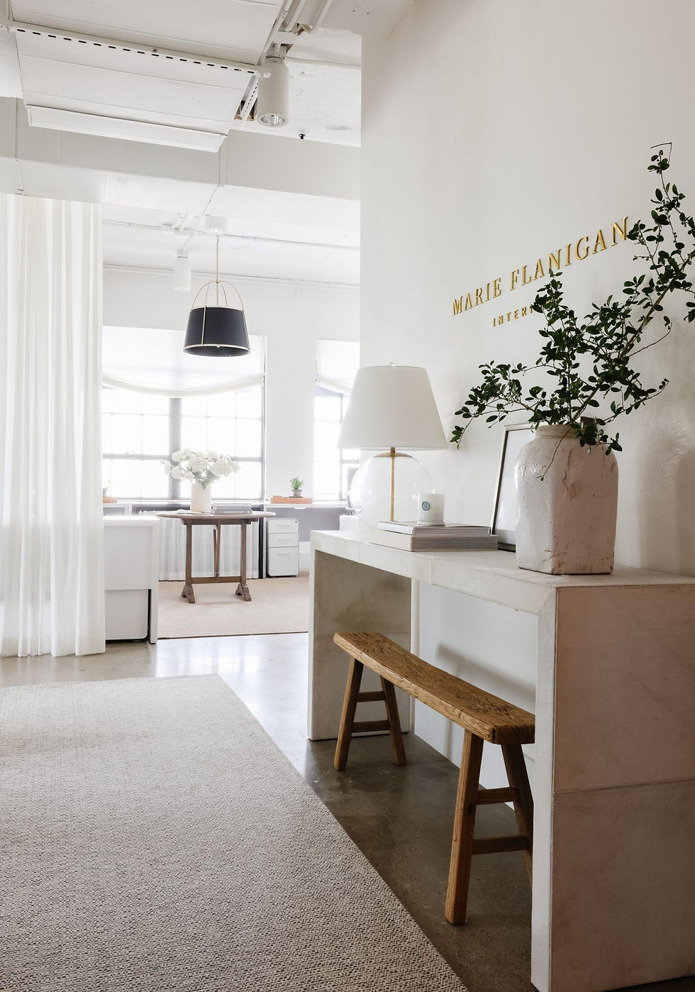 Modern office with white walls, wooden bench, and a plant on a console. Soft lighting, pendant lamp, and "Marie Flanigan Interiors" on wall.