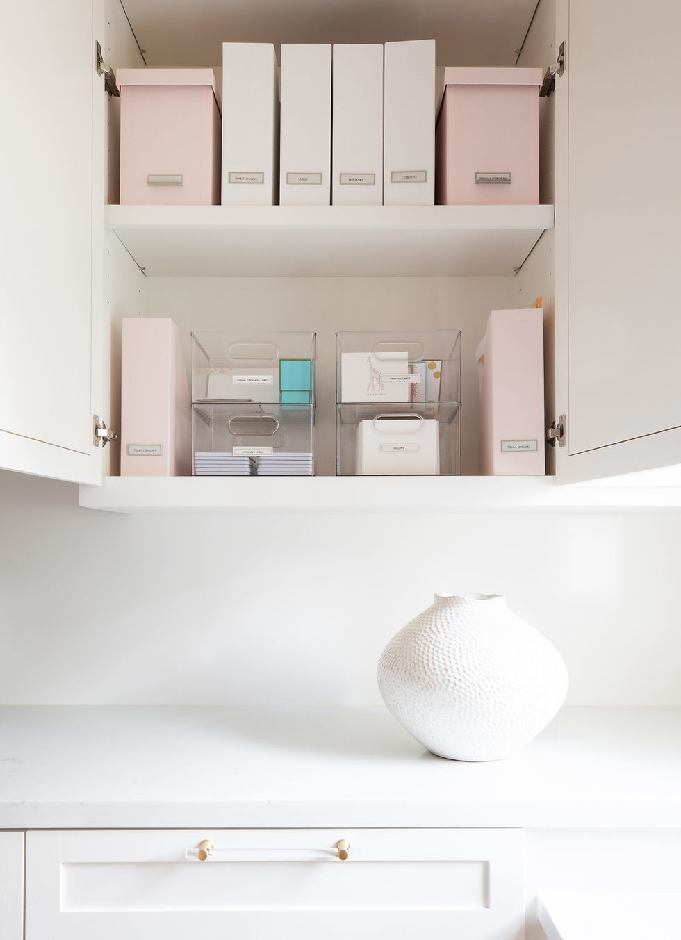 Organized shelves with pink and white boxes, labeled, and clear containers. A textured white vase is on the counter below. Minimalist decor.