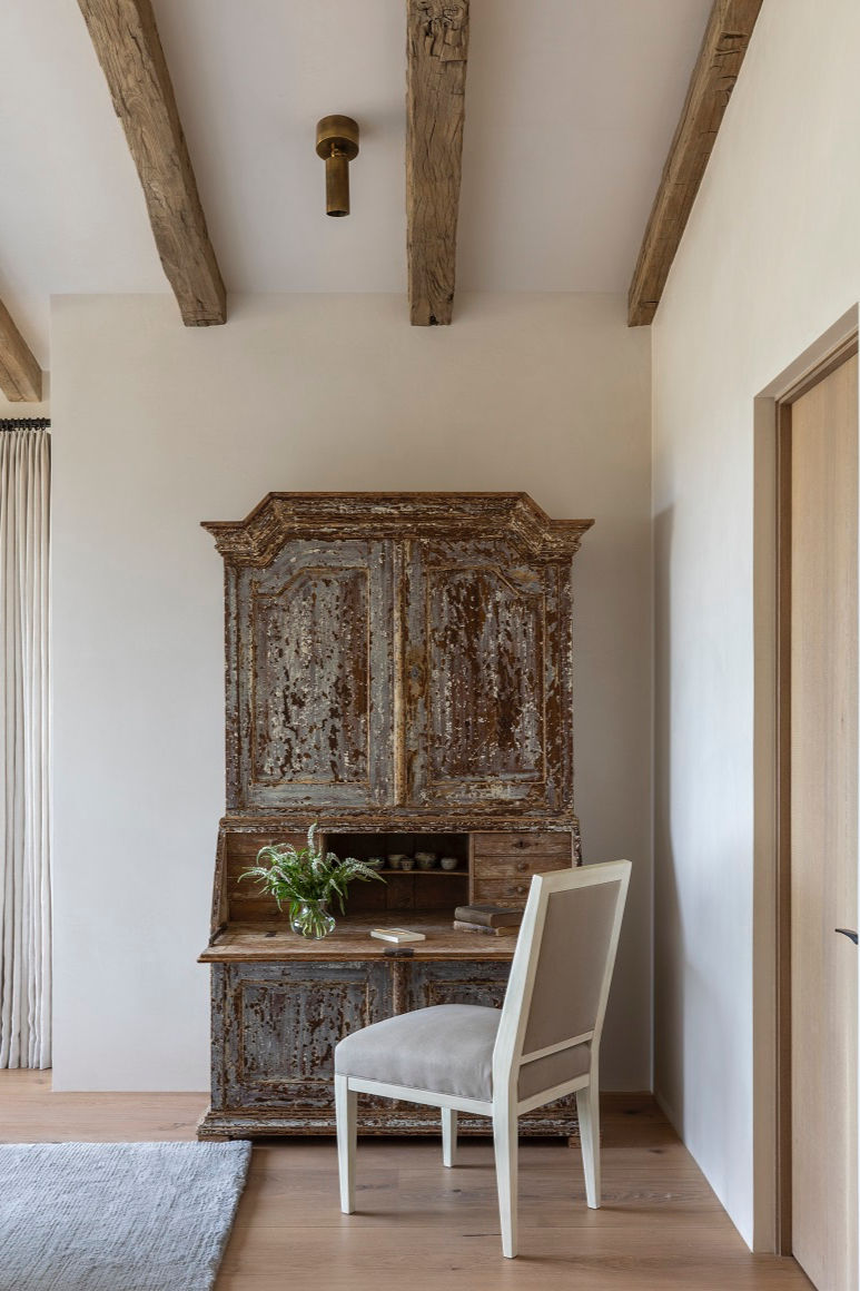 A rustic wooden desk with a distressed finish, a white chair, and a vase with green leaves. Exposed beams on the ceiling in a cozy room.