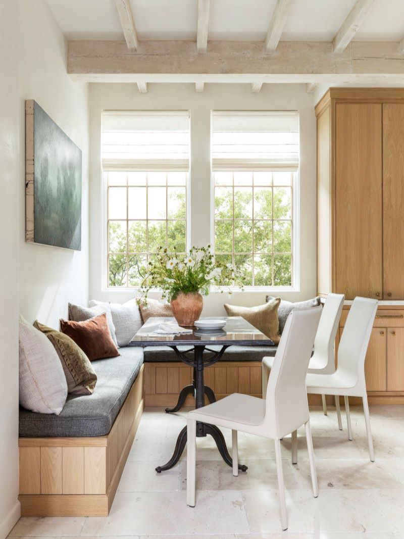 Bright, cozy dining nook with a bench, cushions, white chairs, and a central table topped with a vase of flowers under bright windows.