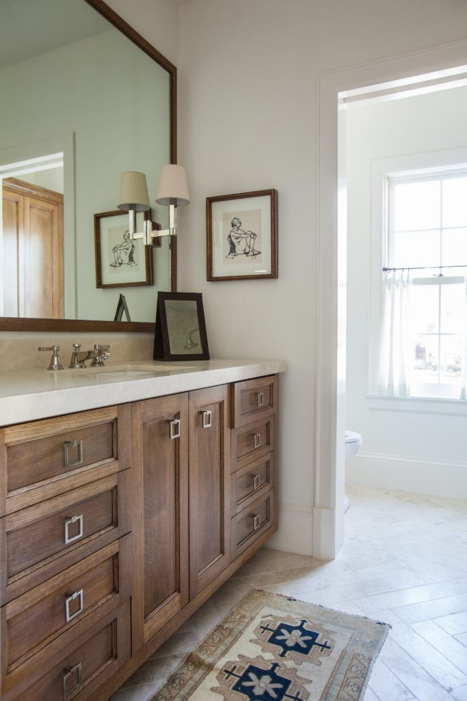 Wooden bathroom vanity with a large mirror, two framed sketches, light fixture, and patterned rug. Bright room with a window view.
