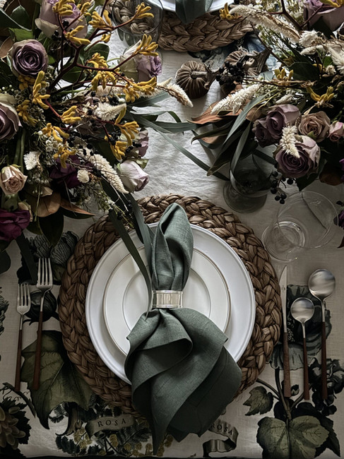 Close-up of a layered place setting with woven charger, green linen napkin, floral table linens, and seasonal blooms