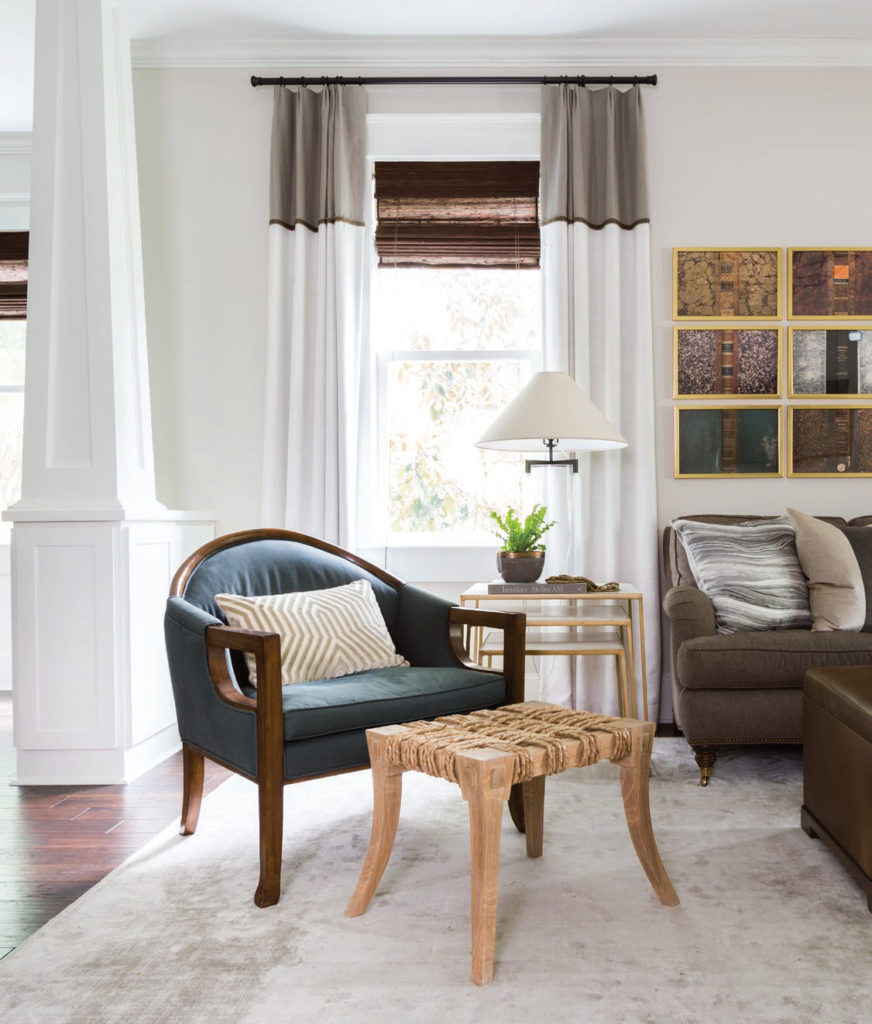 Cozy living room with a blue chair, striped cushion, wicker stool, and side table with a plant and lamp. Neutral tones and patterned art.