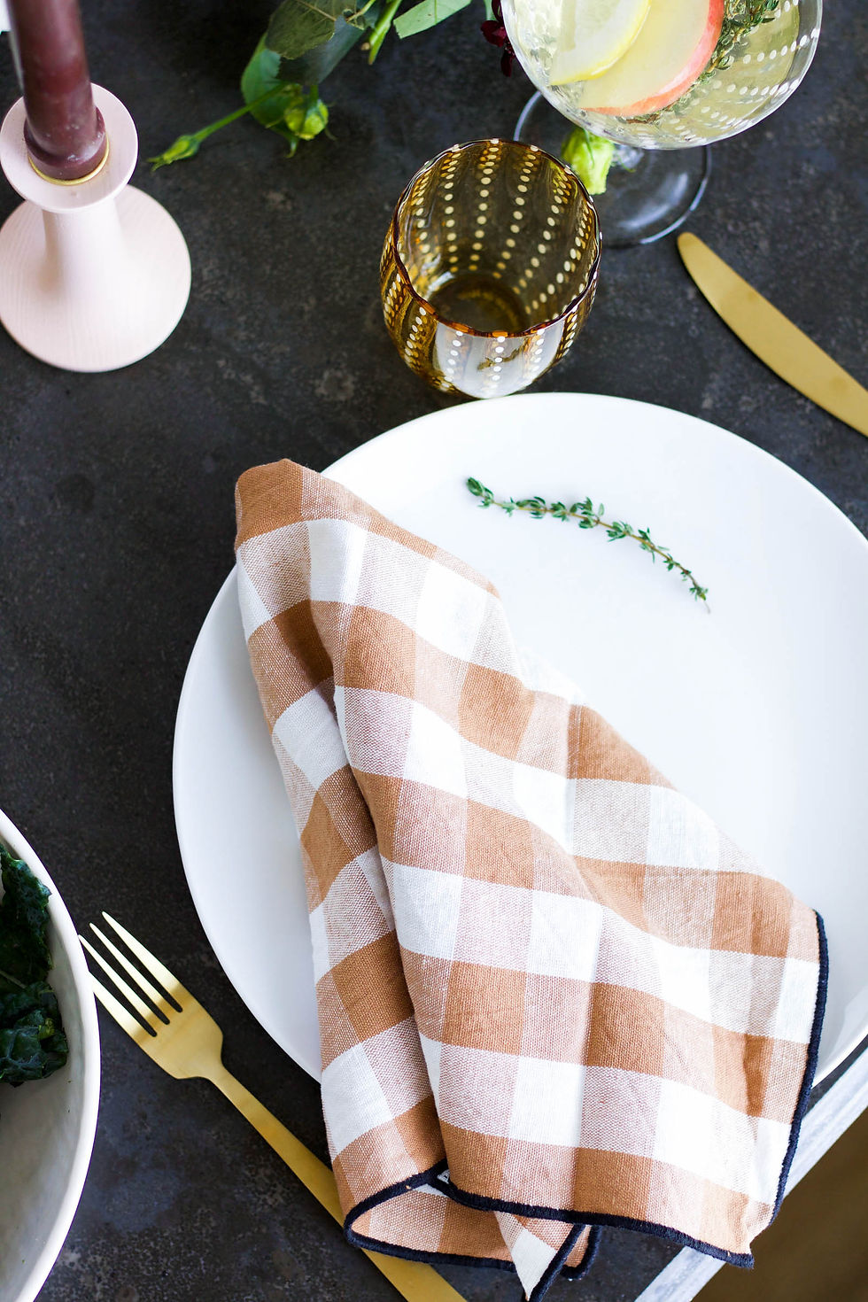Elegant dining setup with a white plate, gingham napkin, gold utensils, dotted glassware, candle, and lemon slice in drink on a dark table.