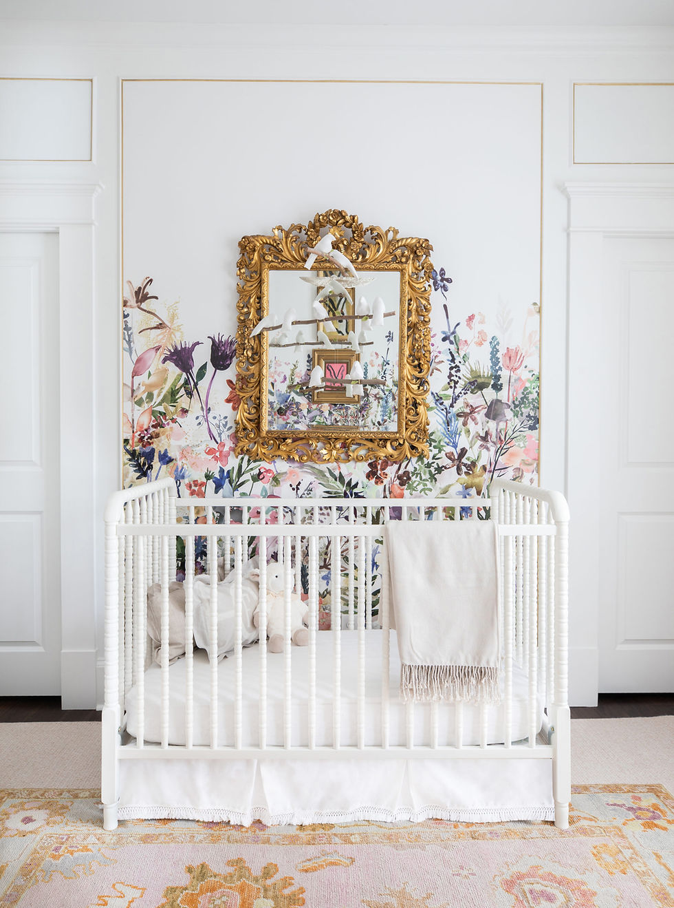 White crib with toys and blanket, set against floral wallpaper. Ornate gold mirror above, bright and peaceful nursery setting.