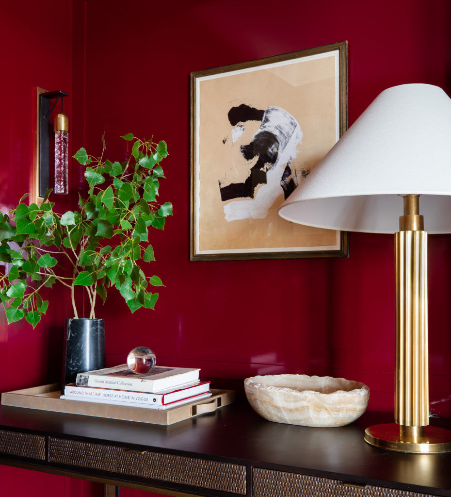 Luxe setting with a gold lamp, leafy plant, art piece, and books on a dark table against a red wall, creating a sophisticated ambiance.