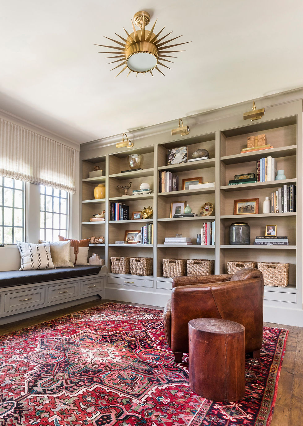 A cozy study features a brown leather chair, a vibrant red patterned rug, bookshelves with decor, and a sunburst light fixture. Natural light filters in.