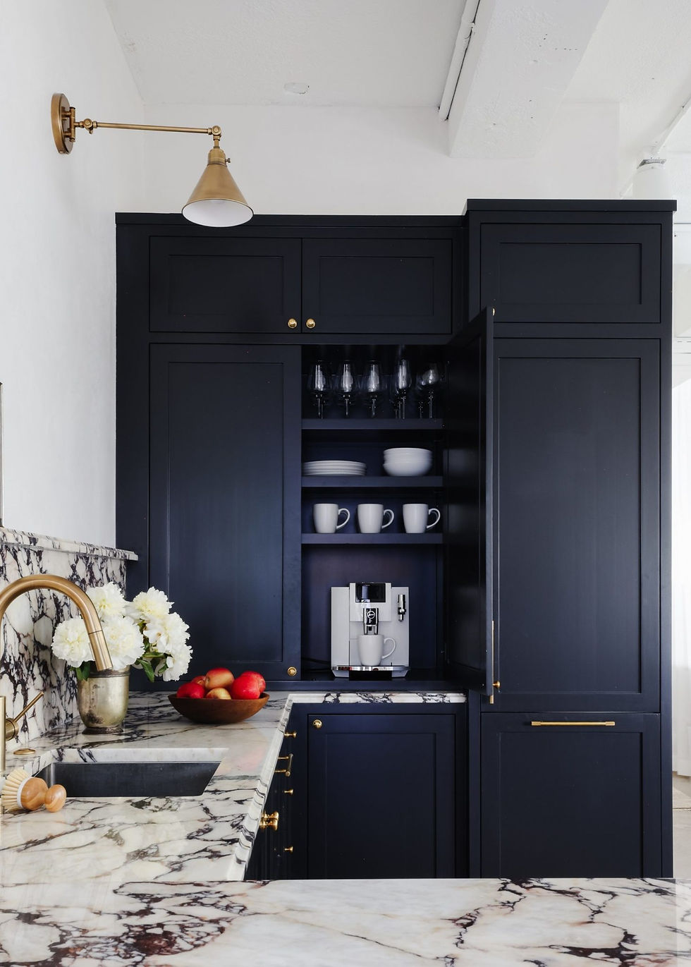 Sleek kitchen with dark cabinets, open to reveal cups and a coffee machine. Marble countertops hold a vase of flowers and apples.