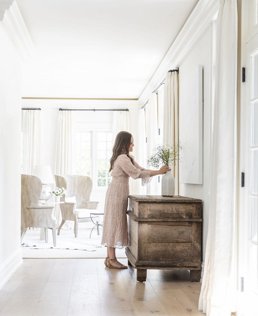 Marie Flanigan arranging flowers in a vase on a rustic wooden chest in a bright, white room. Curtains and light wood floors create a serene mood.