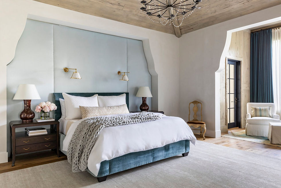 Elegant bedroom with a plush bed, blue accents, lamps, and flowers. A chandelier hangs overhead. A chair and armchair sit near sheer curtains.