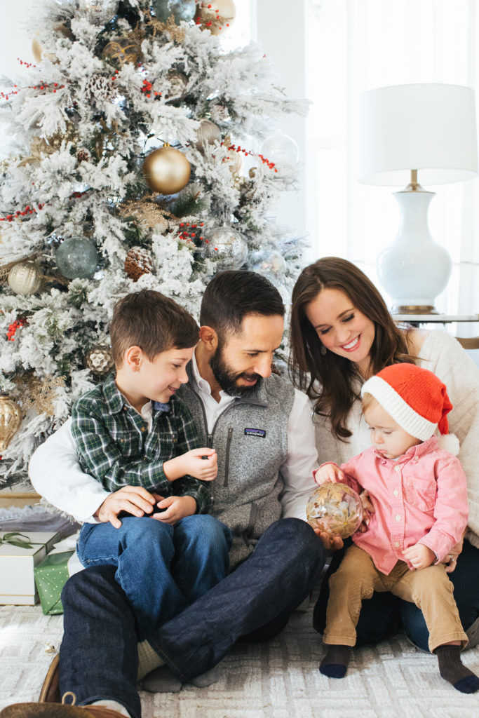 A family of four sits by a snowy Christmas tree. Parents and kids share smiles, with the youngest in a Santa hat, holding a glass ornament.