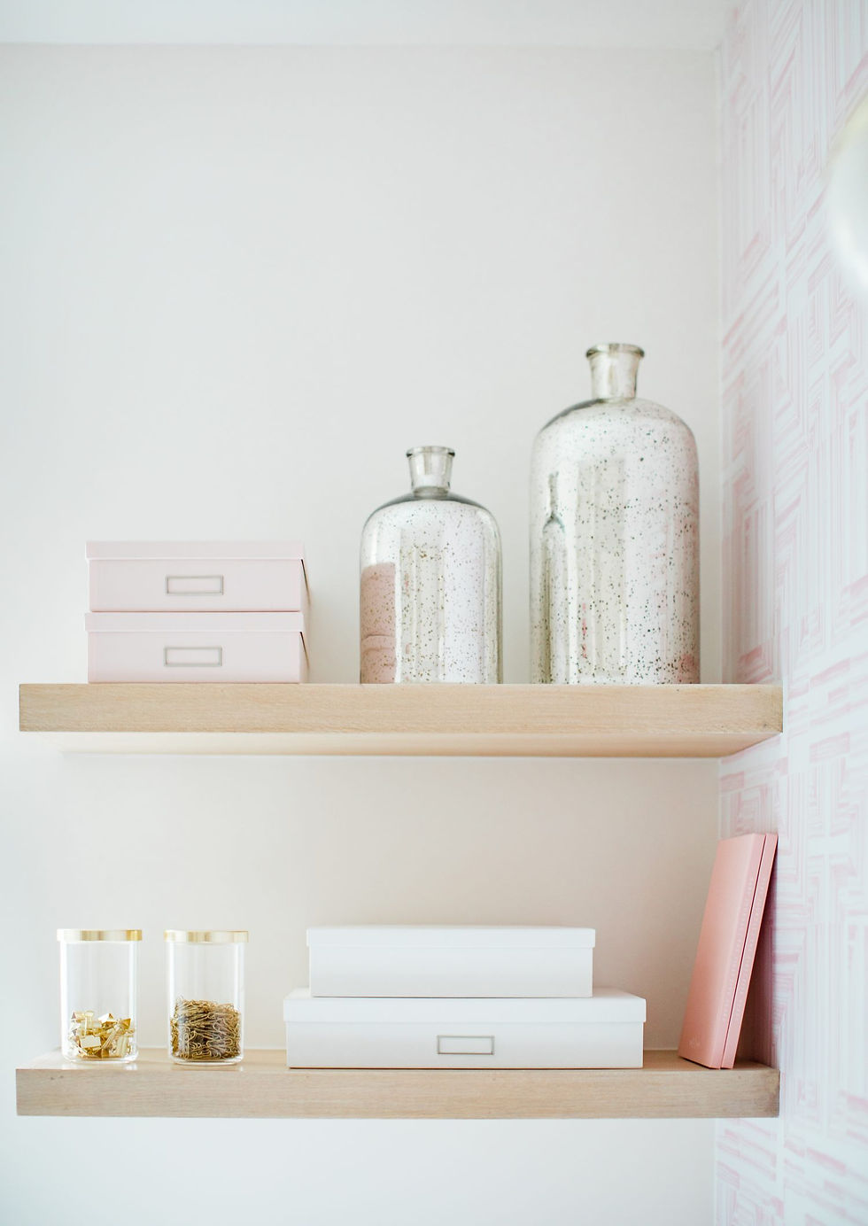 Wooden shelves with decorative glass bottles, pink and white boxes, and glass jars with clips. Soft pink patterned wallpaper backdrop.