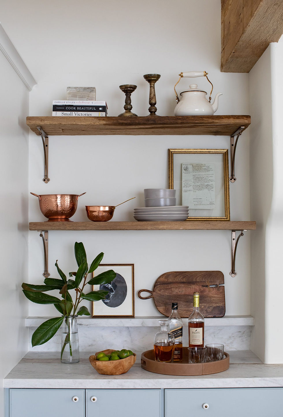 Kitchen shelves with copper bowls, white kettle, and candlesticks. Below, a vase with leaves, framed art, wooden tray, and bottles. Cozy vibe.