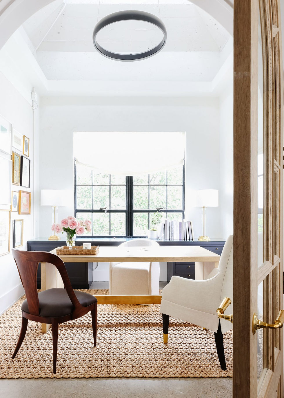 Bright home office with a wooden desk, chairs, and a woven rug. Large window, pink flowers, books, and art on walls. Modern, airy feel.