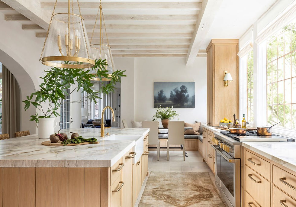 Bright kitchen with light wood cabinets, marble countertops, gold fixtures, and green plants. Large windows and cozy seating area with art.
