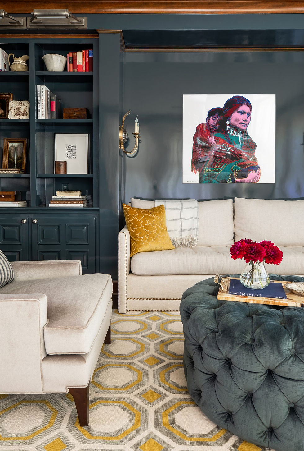 Cozy living room with beige sofa, mustard pillow, blue ottoman, and geometric rug. Colorful art on navy wall. Vase with red flowers.