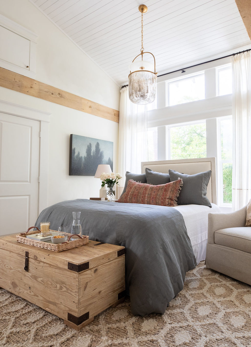 Cozy bedroom with a gray bed, decorative pillows, wooden chest, and tray. Bright windows, white walls, and pendant light enhance elegance.