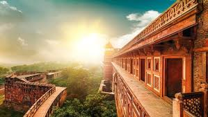 Sunset illuminates a historic red stone fort with intricate carvings, lush greenery below, and a serene sky above; evokes tranquility.