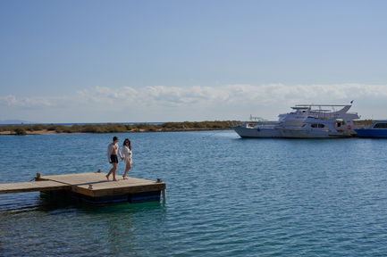 Couple walking on a jetty be the Red Sea, with big boat in the background and Mangroove trees 