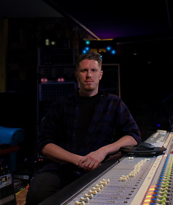 Luke is sitting behind a mixing console, facing the camera. He is wearing black jeans and a dark flannel shirt. His hair is short with a fade on the sides. The background is dark with a purple hue and blurred to focus on Luke.