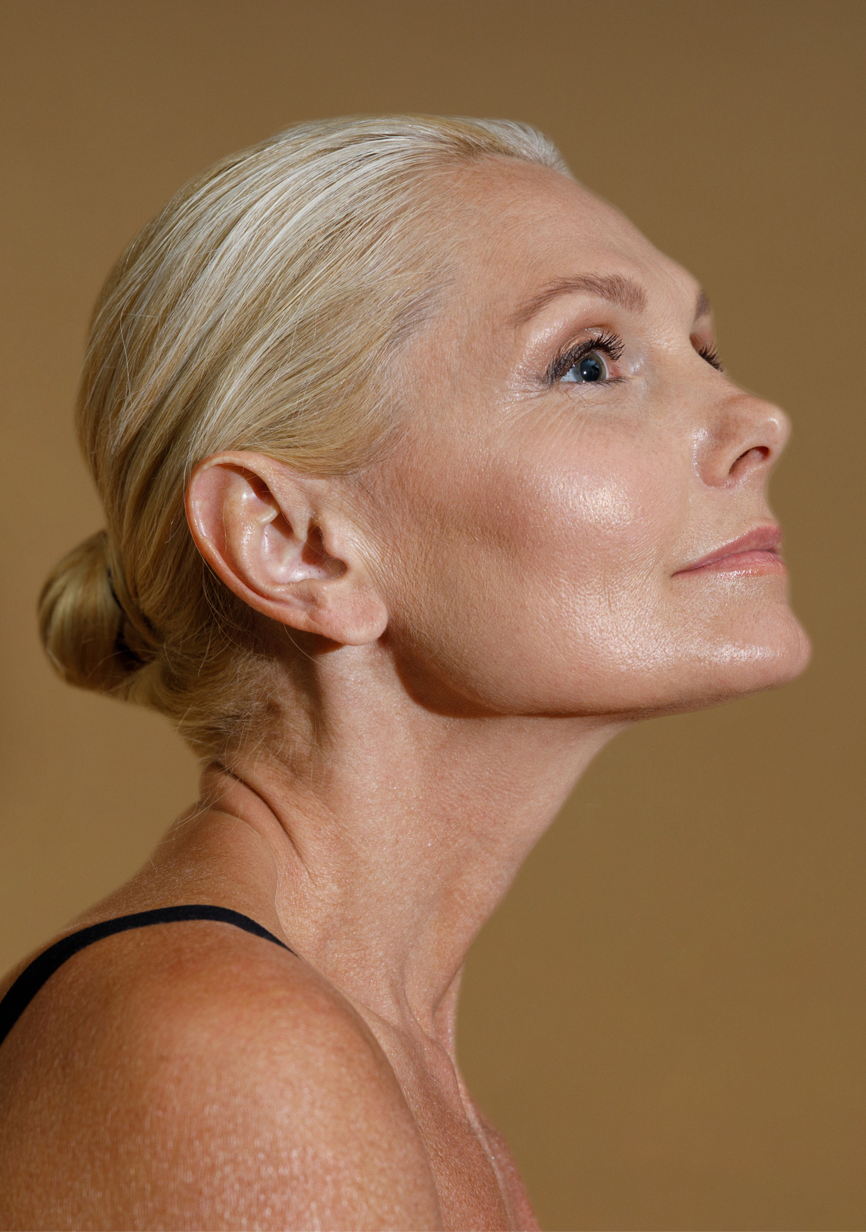 Profile portrait of a blonde woman with hair in a bun, gazing upward against a warm beige background, calm and poised.