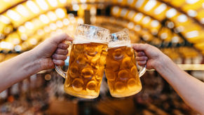Two hands clink large frosty beer mugs in a warm, blurred beer hall with golden lights.