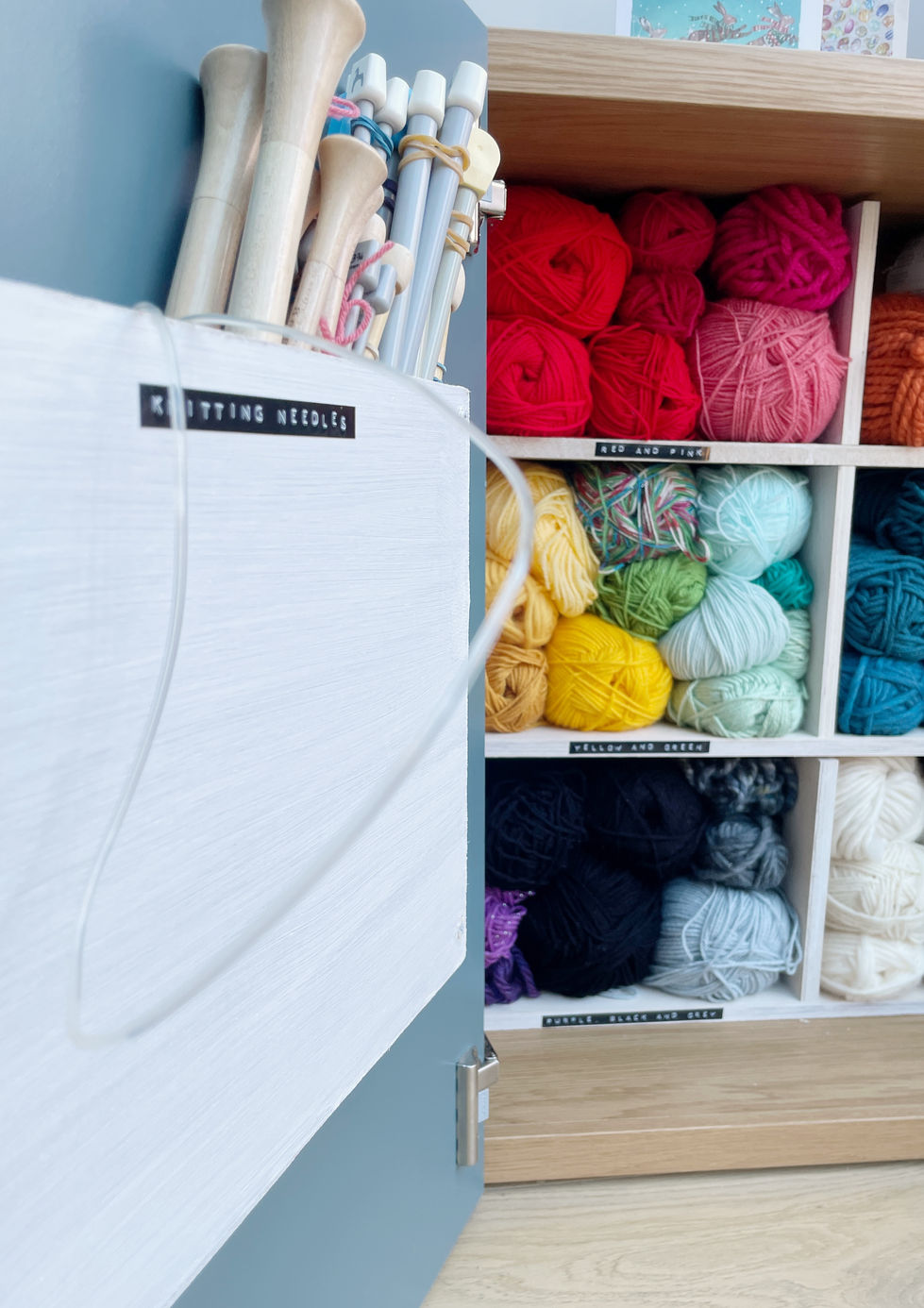 A knitting cupboard is shown organised and tidied. In the foreground is a custom built box mounted onto the inside of the door with a label on it reading ‘knitting needles’. Paired up knitting needles poke out of the top. In the background colourful balls of yarn are organised by colour into custom built cubbies, all labelled with a retro embossed label.