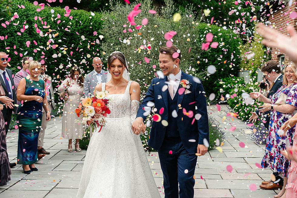 Bride in lace gown and groom in navy suit walk hand in hand under colorful confetti, surrounded by joyful guests in a garden setting.