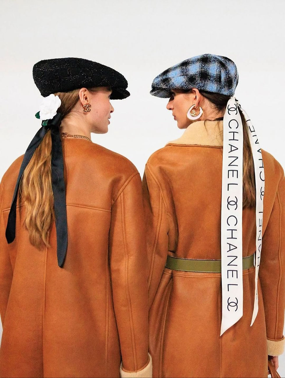Two women in brown coats face each other wearing hats; one black and one blue plaid. A white ribbon with "Chanel" text is visible.