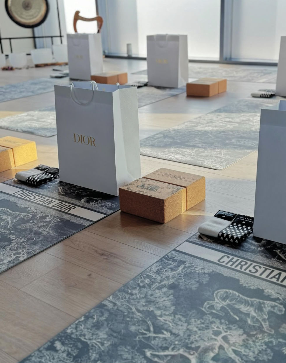 Yoga mats with "Christian Dior" text, cork blocks, and Dior gift bags arranged neatly on a wooden floor in a bright room.