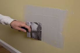 Hand repairing a wall using drywall compound and a putty knife on a beige wall. The patch is slightly lighter, creating a contrast.