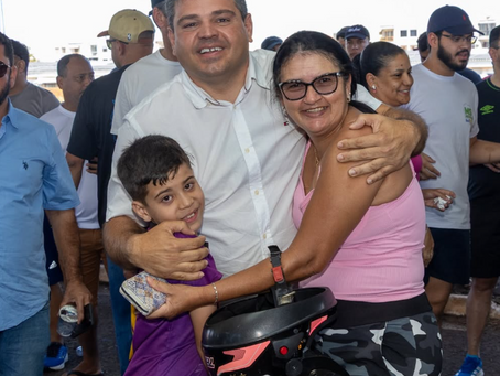 Silvio Pitu pré-candidato a deputado estadual em Mato Grosso do Sul abraça mulher e criança sorridentes em evento ao ar livre, com multidão ao fundo; ele de branco, ela de rosa.