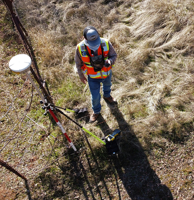 An overhead view of a surveyor in a high-visibility vest and blue hard hat walking through dry, rugged brush while operating a surveying data collector pole.