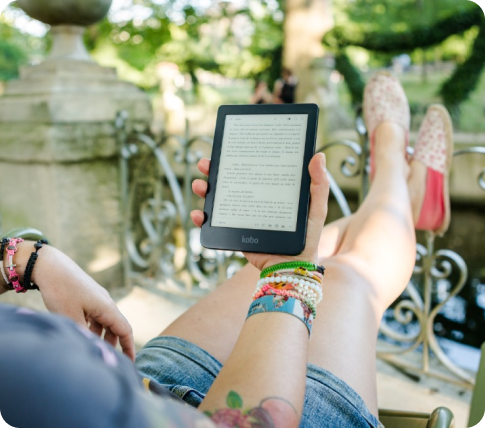 A person sitting on a bench with crossed legs, holding an e-reader with text on the screen, in a park-like setting with greenery and sunlight.