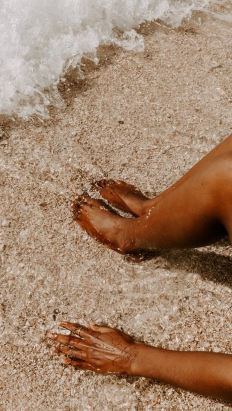 Tanned feet and hands at the water’s edge on sandy beach.