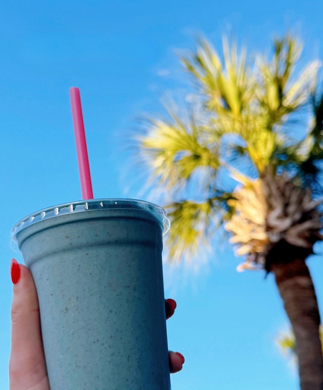 Aqua-blue smoothie cup with pink straw held up to palm trees.