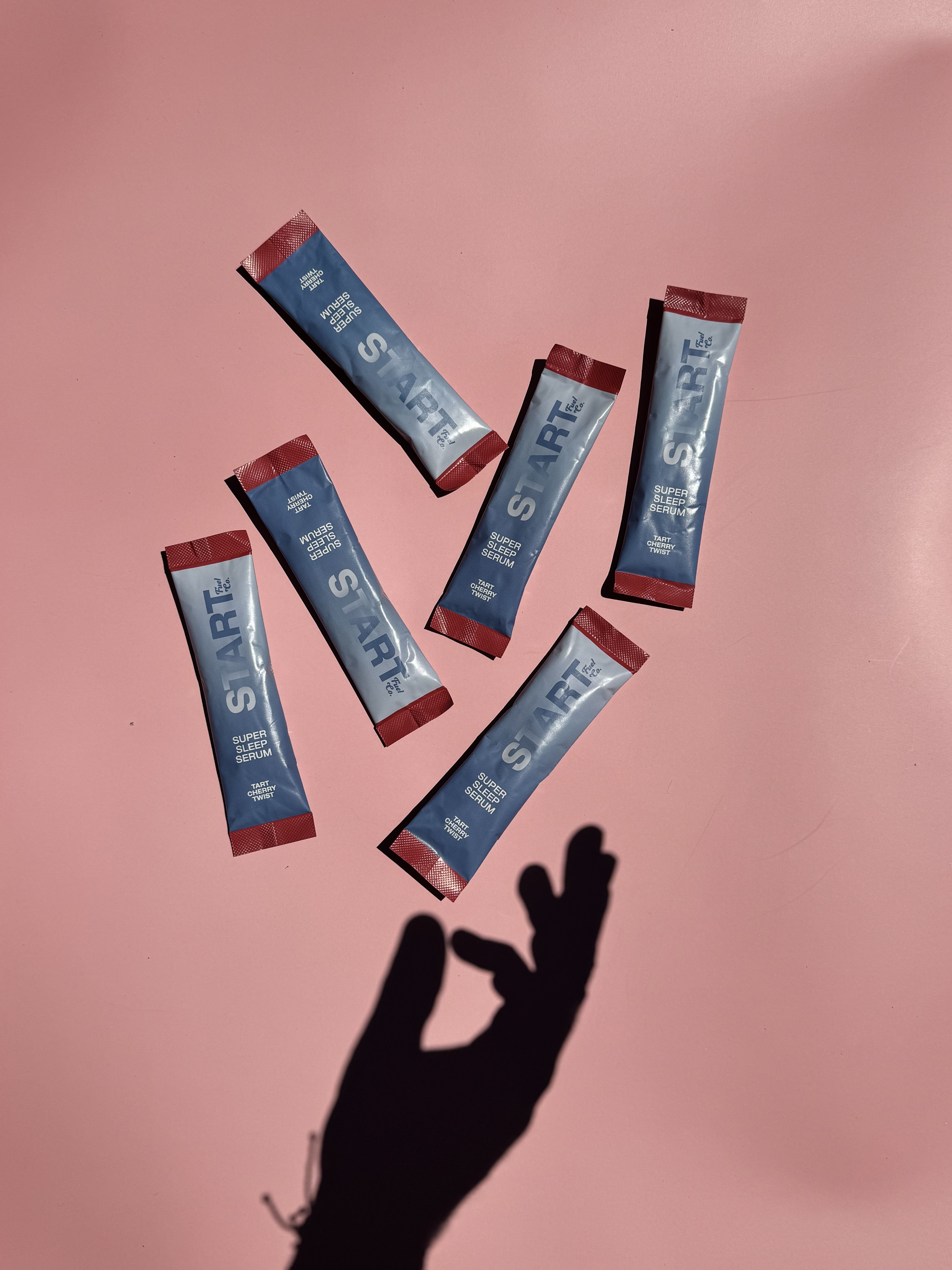 Hand reaching toward scattered Super Sleep Serum stick packs on a pink background.
