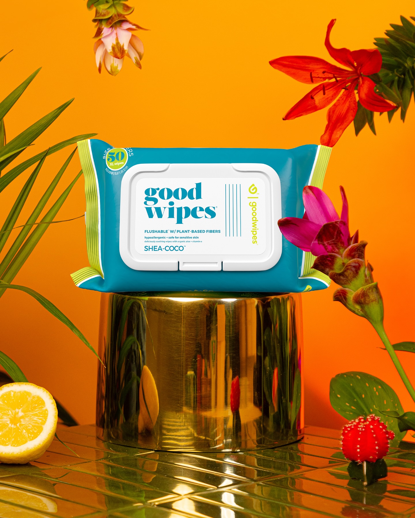 Goodwipes pouch on gold pedestal with flowers and lemon against orange backdrop.