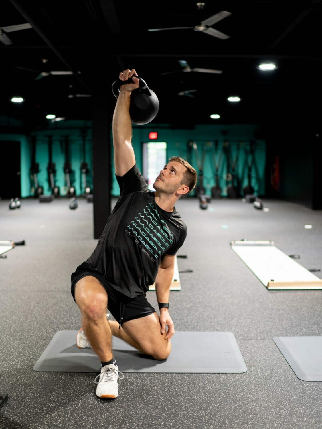 Trainer performs overhead kettlebell reverse lunge on turf while cueing class at MADabolic Atlanta.