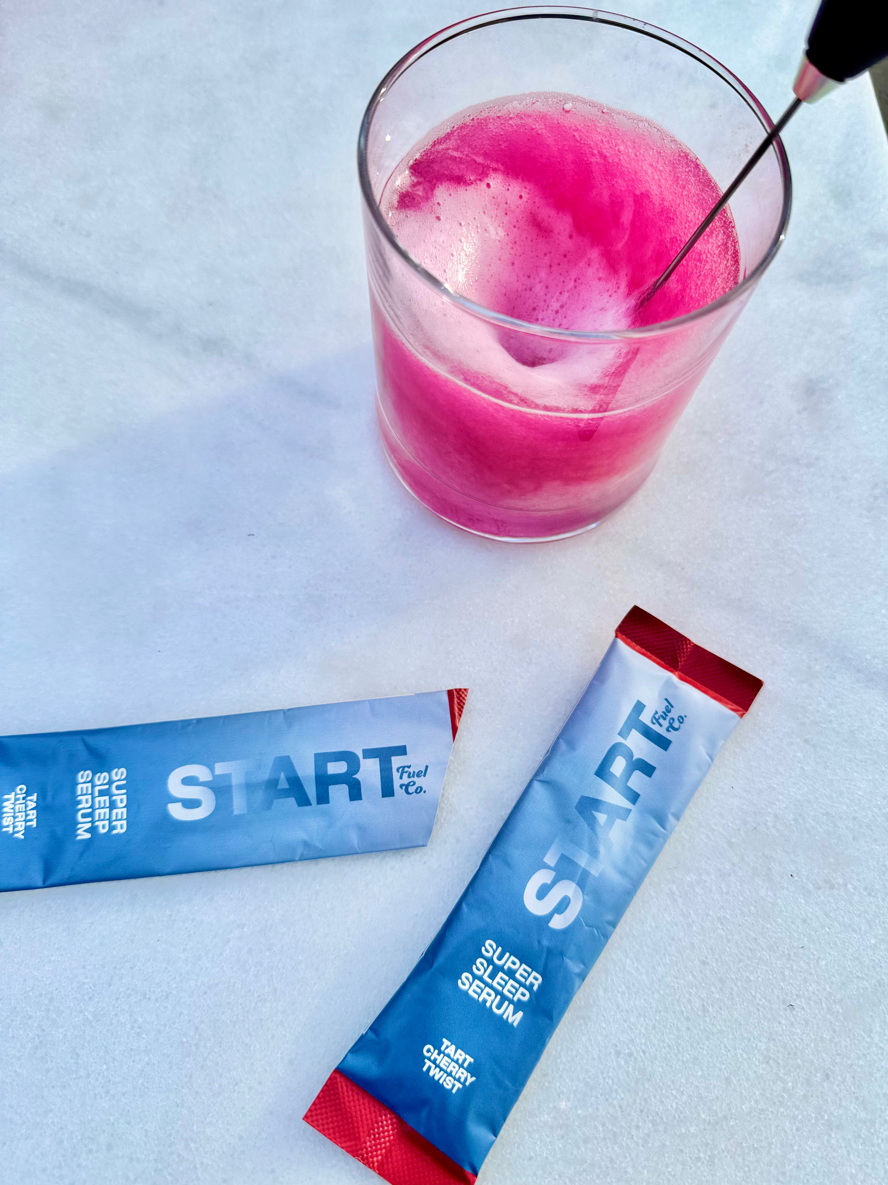 Overhead of pink beverage with cherry and two Super Sleep Serum sticks on a marble surface.