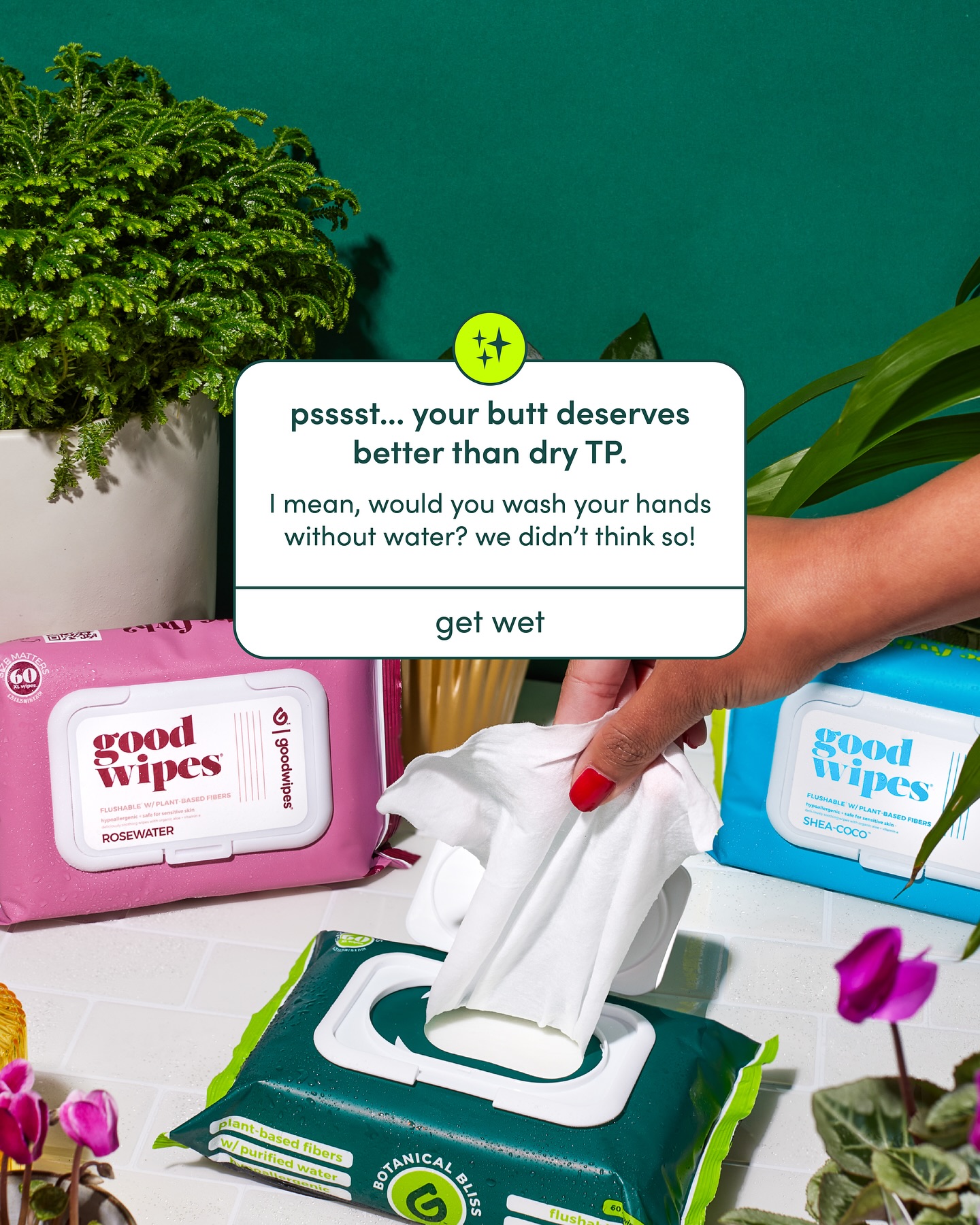 Hand removing a Goodwipes wipe from a pack resting on grass with plants nearby.